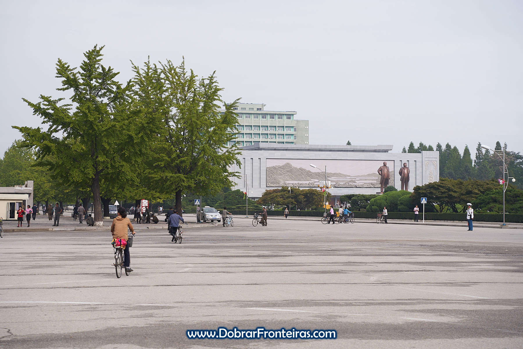 Pessoas de bicicleta em rua da Coreia do Norte com estátuas dos líderes ao fundo